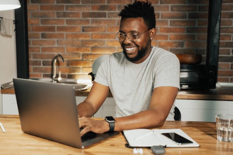A man with a cheerful expression sits at a table, engrossed in his work on a laptop.